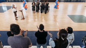 Graduates standing and clapping in a gym after receiving diplomas during a graduation ceremony.